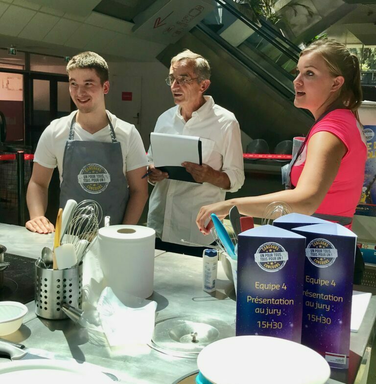 Concours culinaire, deux candidats et un juré autour d'un plan de travail