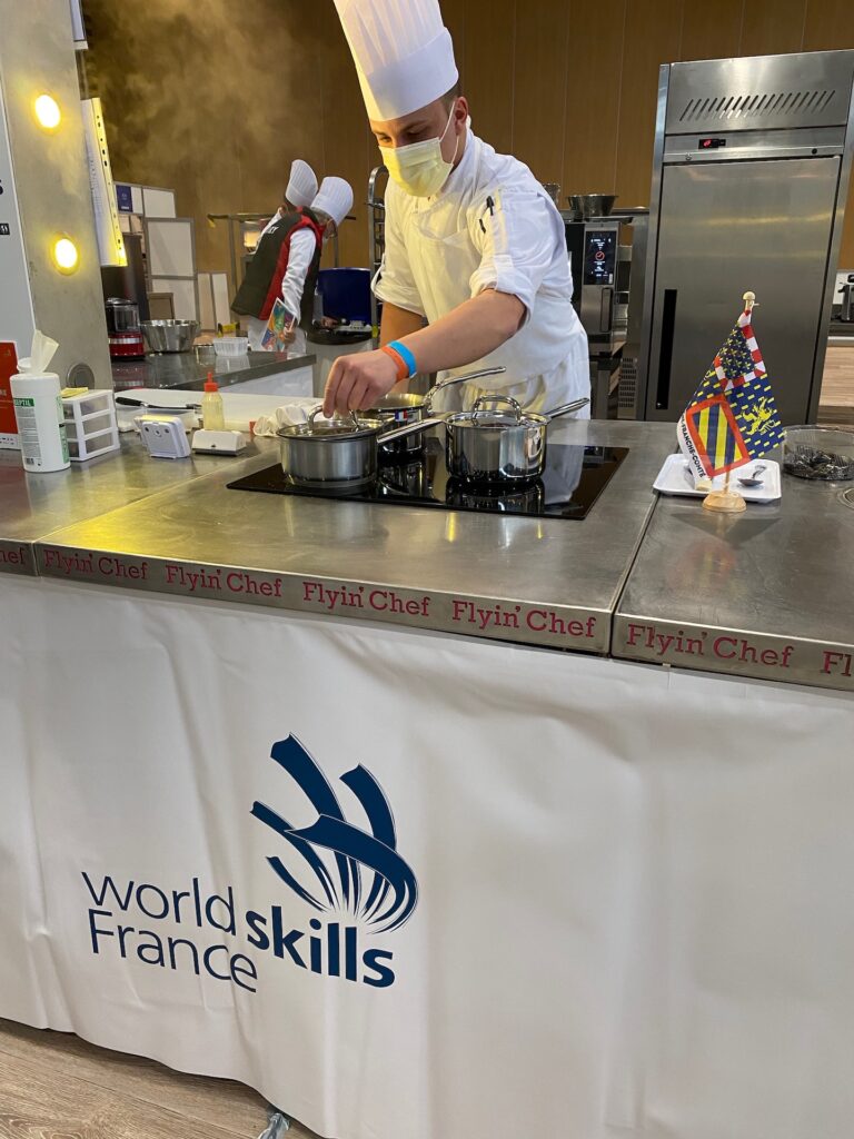 Candidat de la finale nationale de Lyon en pleine cuisson sur un poste habillé WorldSkills France, avec le fanion de sa région