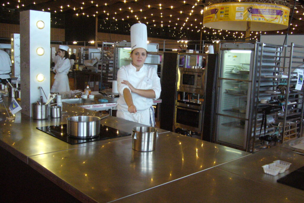 Candidate en toque à son poste de cuisine sous la bannière Olympiades des Métiers et les guirlandes lumineuses