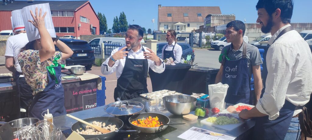 Chef expliquant une recette à des participants en tablier personnalisé, lors d'un atelier culinaire participatif Flyin'Chef en plein air